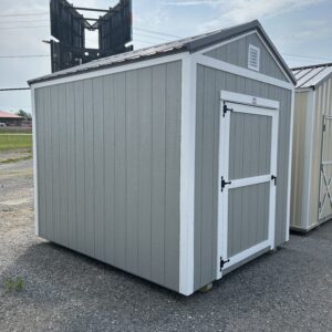Gray storage shed with white trim on a gravel lot, with a large metal sign structure in the background.