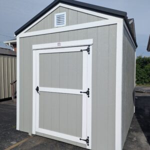 Grey portable storage shed with white trim, a large framed door with black hinges, and a small vent near the peak of the roof on a paved surface.