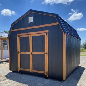 Dark gray wooden shed with tan trim and double doors on a gravel lot under a blue sky with clouds.